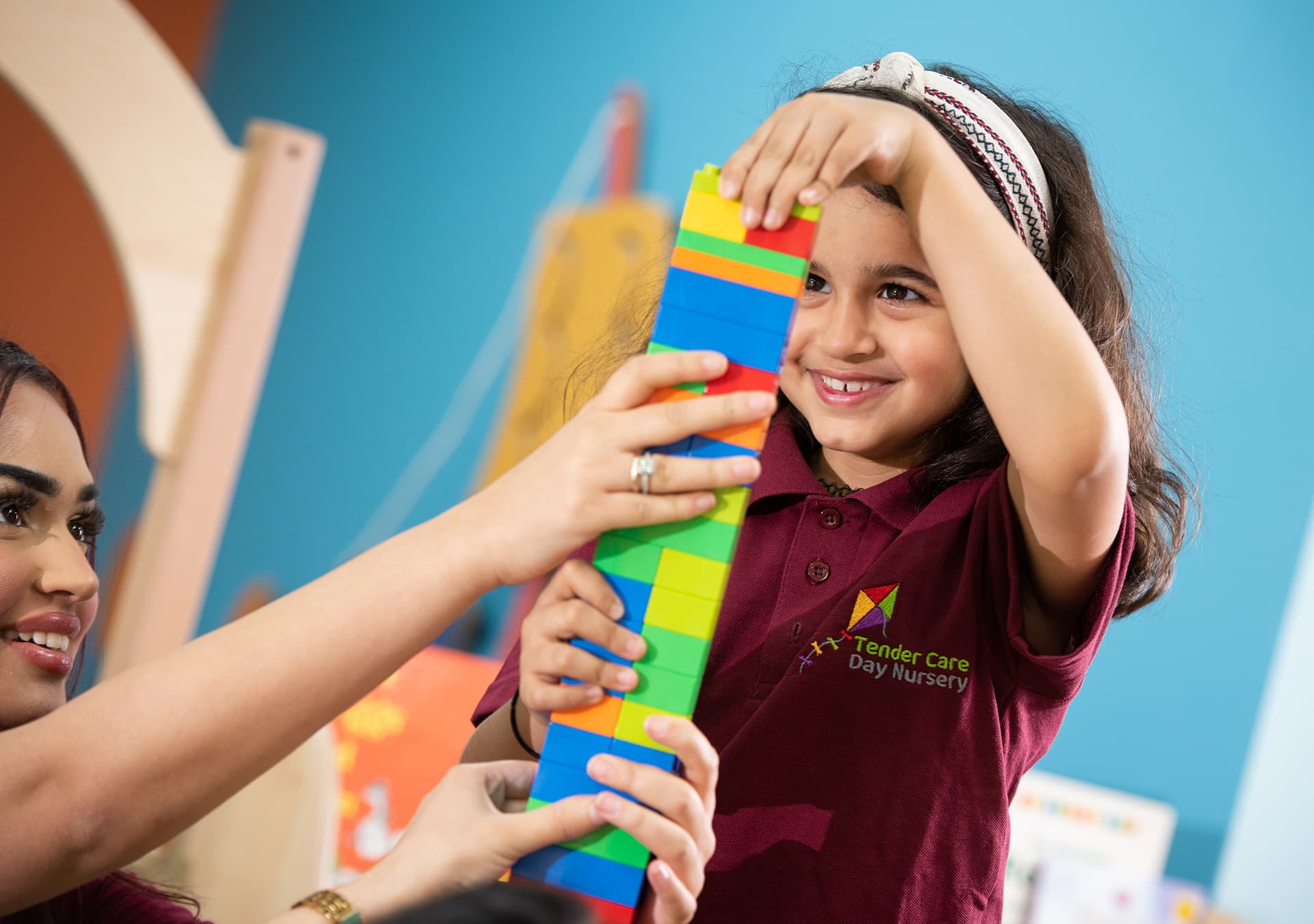 Smiling girl building a tall tower of colourful building blocks with a staff member