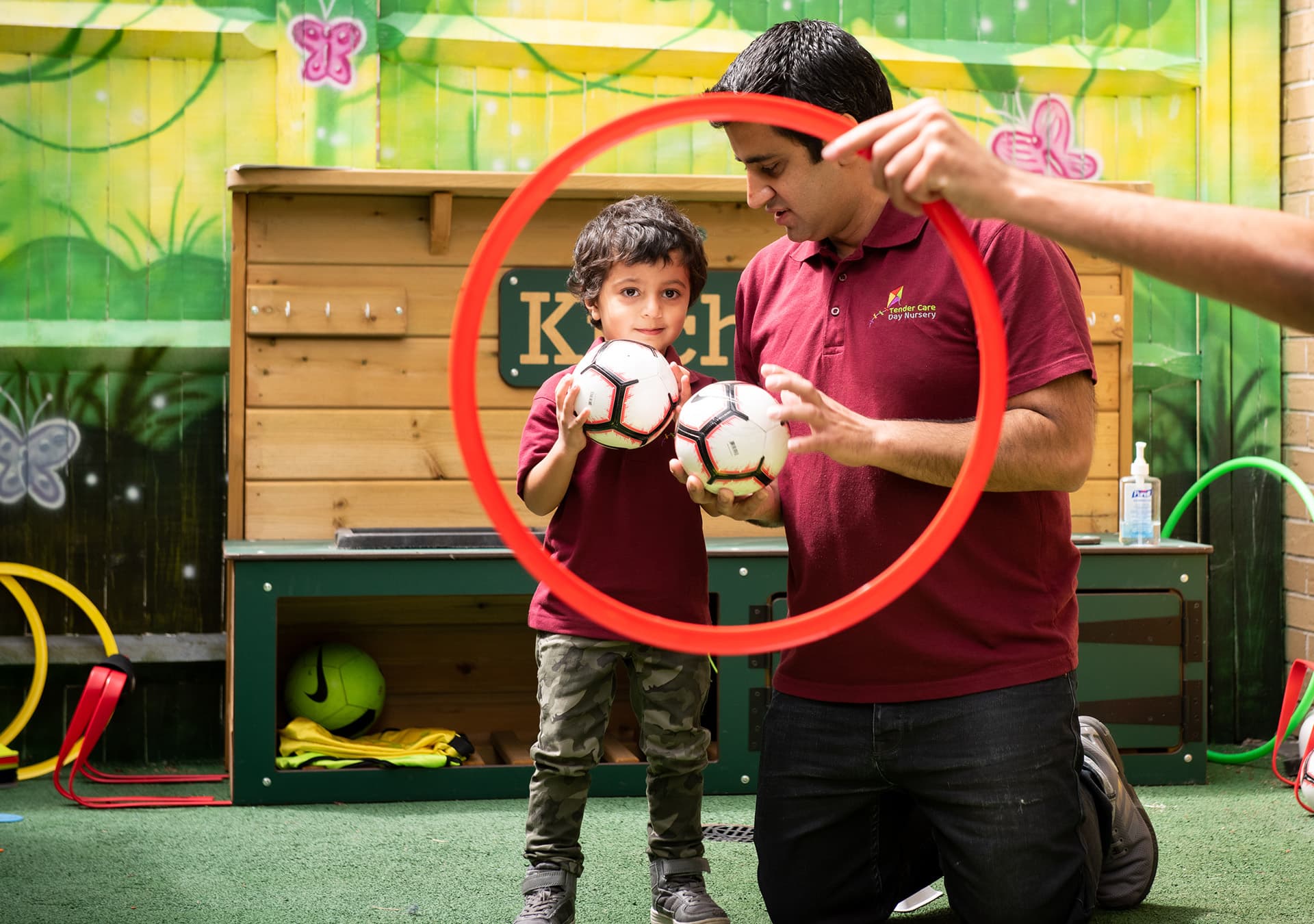 Nursery worker helping a young boy hold two footballs inside a red hoop
